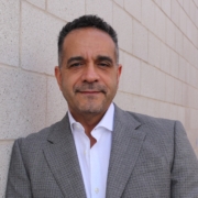 John Vicente in white oxford shirt and gray blazer against off-white brick wall