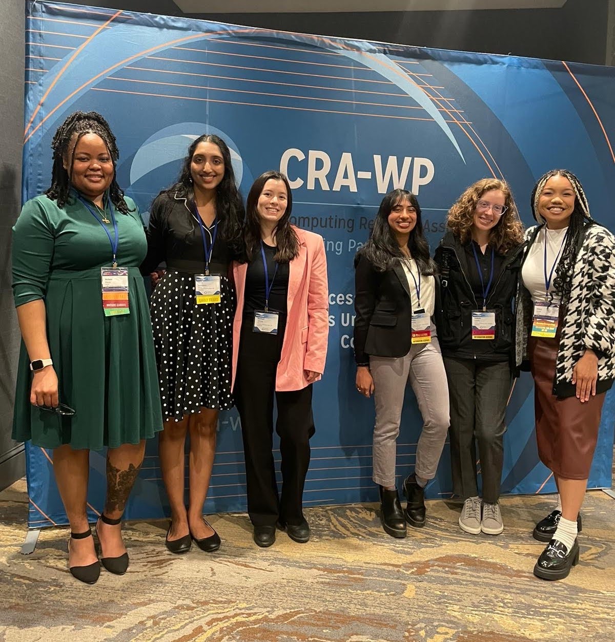 six women wearing conference badges standing in front of a blue CRA-WP banner at the CRA-WP Grad Cohort for Women Workshop