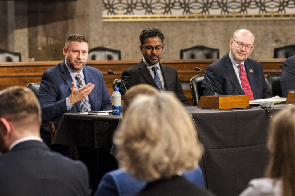 While sitting at a table, Gary Givental, left side of three people, gestures while making his remarks. Dr. Balaprakash sits to his immediate left, followed vy Dr. Reed.
